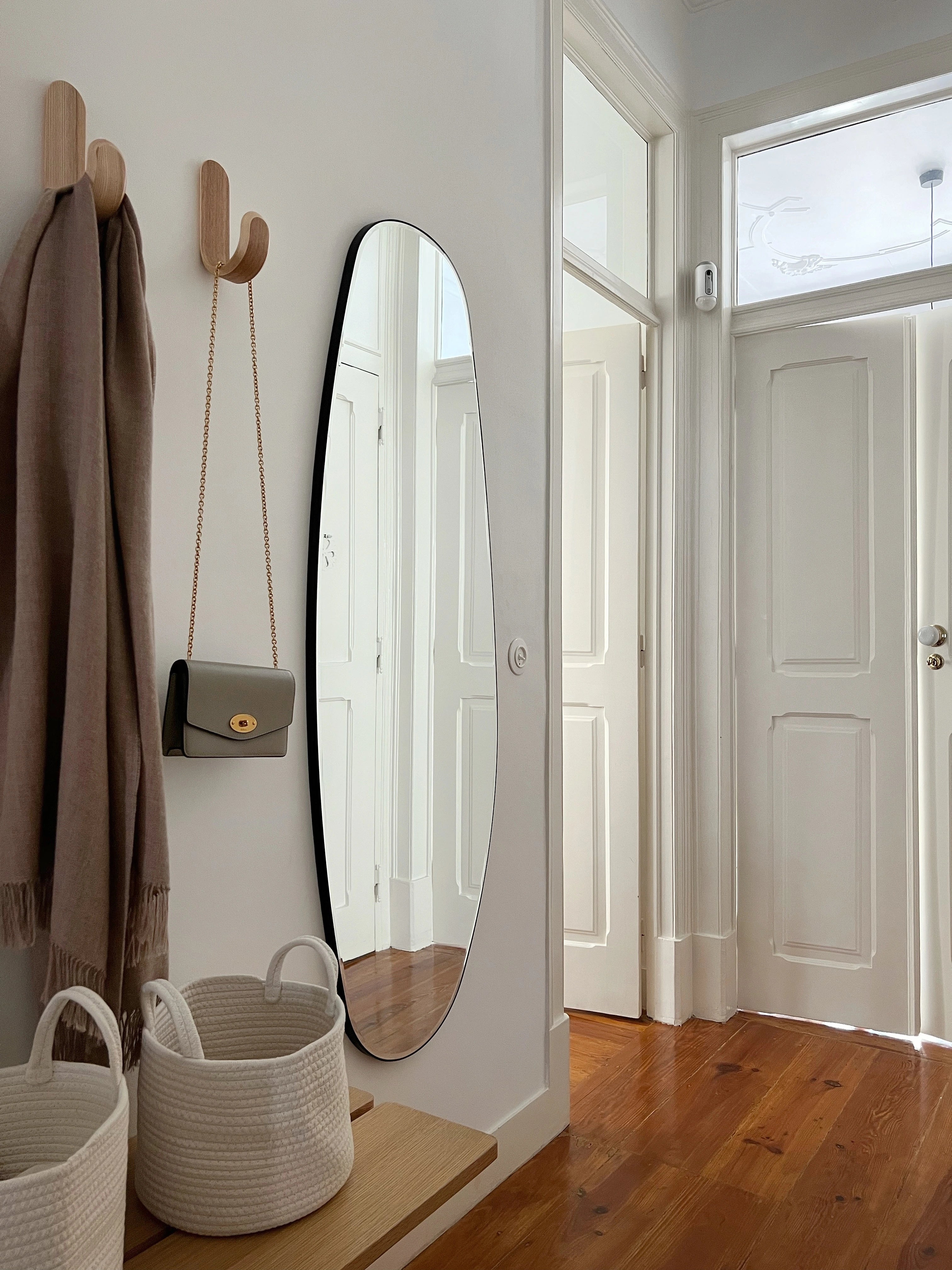 A bright hallway with white walls, wooden floor, a tall opal mirror, two wooden hooks holding a beige scarf and small purse, and two white woven baskets on a wooden bench—perfect for showcasing your unique decor style.