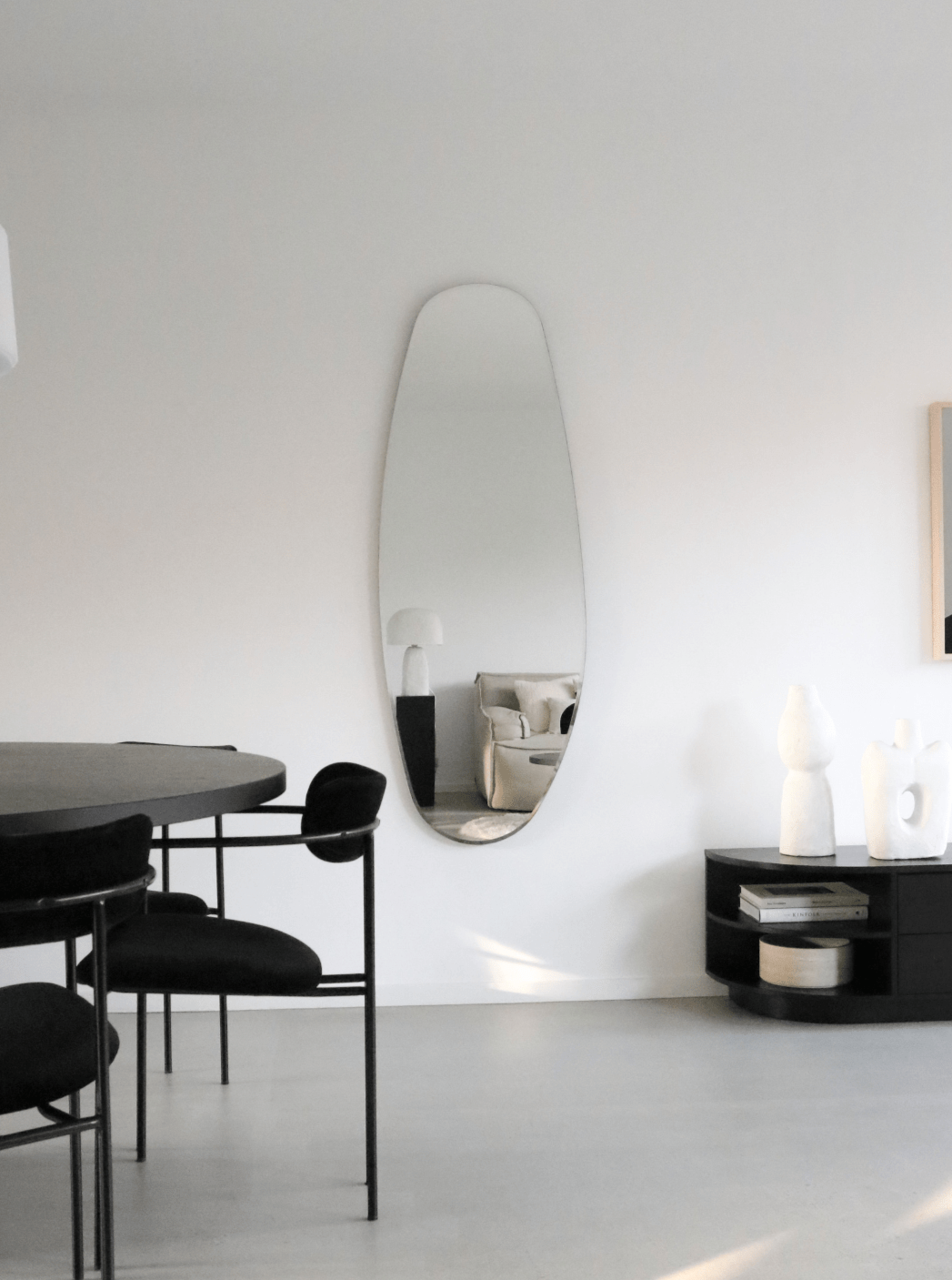 Minimalist living space with a Blossholm opal mirror on a white wall, reflecting a beige sofa and lamp. Black dining chairs, a round table, and abstract white decor pieces complete the unique decor in this modern, bright room.