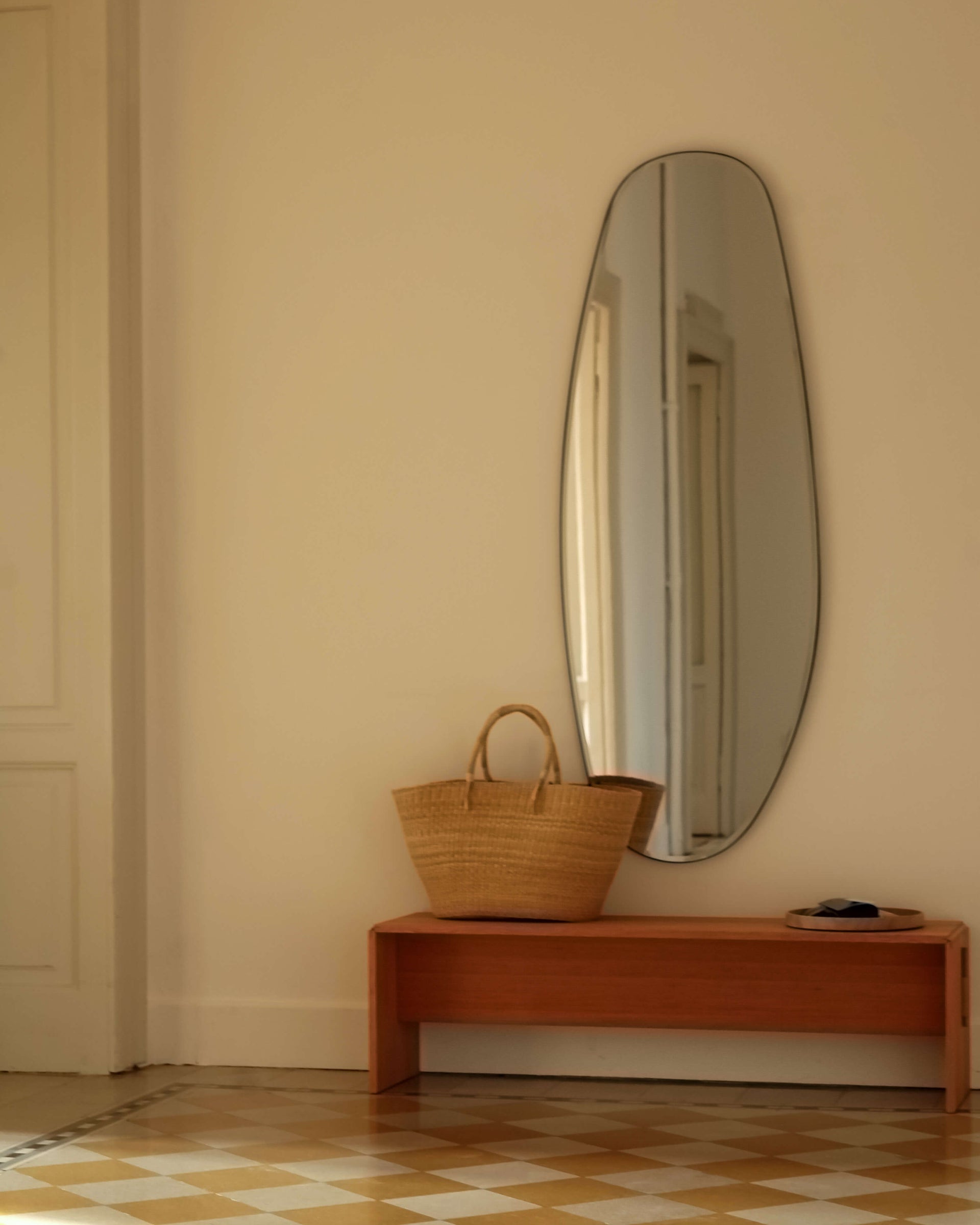 Wooden bench with a woven basket and a large oval mirror on a beige wall.